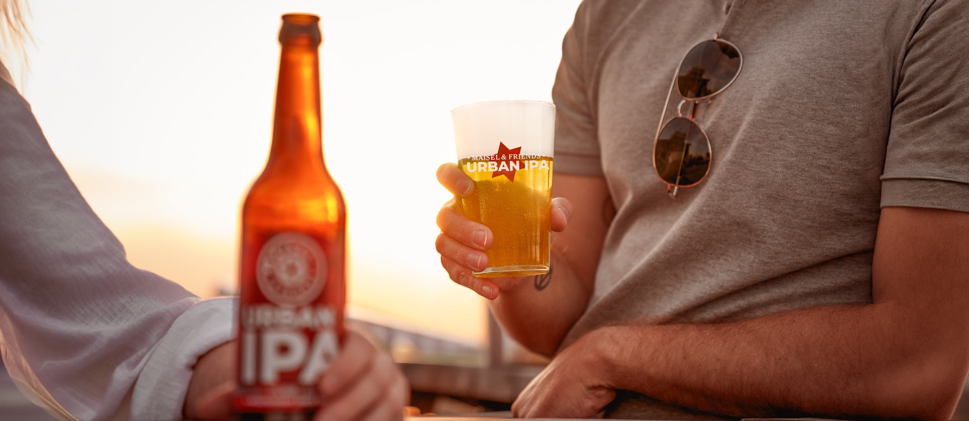 Eine Flasche Urban IPA steht auf einem Tresen und wird von einer Person gegriffen. EIne andere Person hält ein Urban IPA Glas in der Hand, Es ist eine ssommerliche Abendstimmung mit goldenem Licht und Sonnenuntergang im Hintergrund zu sehen.