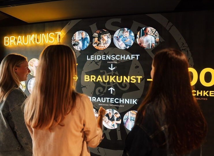 Besucher von Maisel & Friends betrachten einen Bildschirm mit Erklärungen zur Braukunstwelt