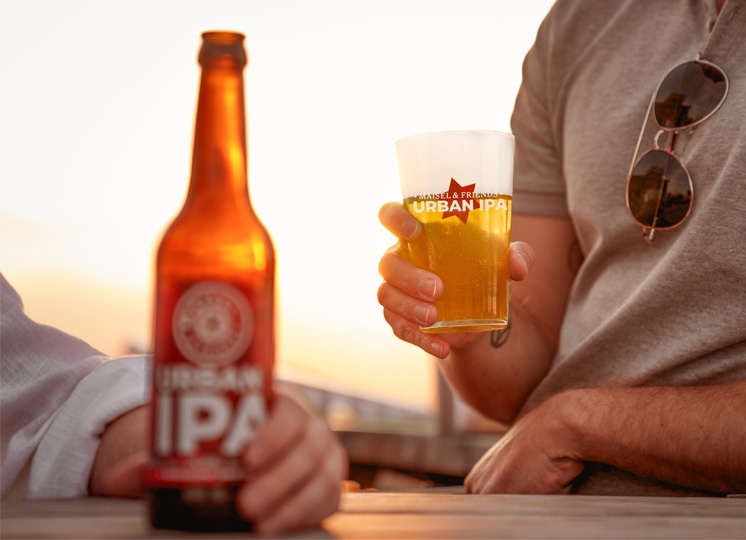 Arbeiten beim Vertrieb von Maisel & Friends in Bayreuth – eine Flasche Urban IPA steht auf einem Tresen und wird von einer Person gegriffen. EIne andere Person hält ein Urban IPA Glas in der Hand, Es ist eine ssommerliche Abendstimmung mit goldenem Licht und Sonnenuntergang im Hintergrund zu sehen.