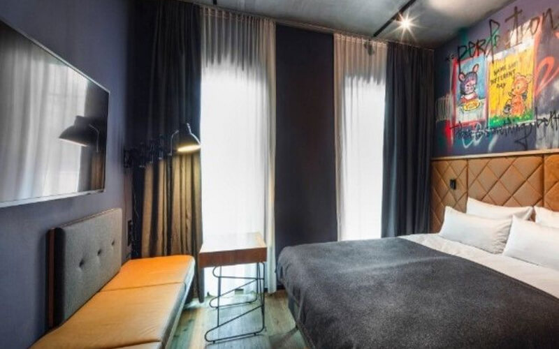 Modernes Hotelzimmer mit Doppelbett, Sitzbank und großer Fensterfront und Wandkunst im Graffiti-Stil