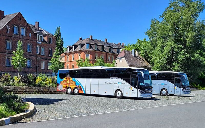 Zwei moderne Reisebusse stehen auf einem Parkplatz vor historischen Gebäuden und Bäumen. Die Szene zeigt die Anreise einer Gruppe zu einem gemeinsamen Ausflug oder Event.