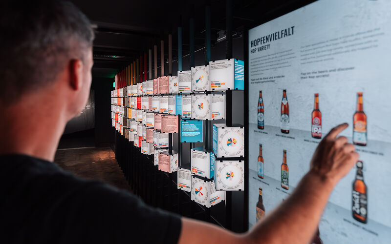 Eine Person steht vor einer interaktiven Ausstellung und tippt auf ein Display mit Informationen zu verschiedenen Biersorten und Hopfenarten. Die Szene zeigt eine informative Station im Rahmen einer Gruppenführung.